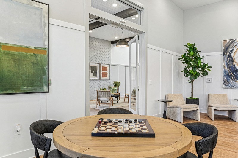 A round wooden table with a checkerboard in the middle is surrounded by chairs in a room with a painting on the wall.