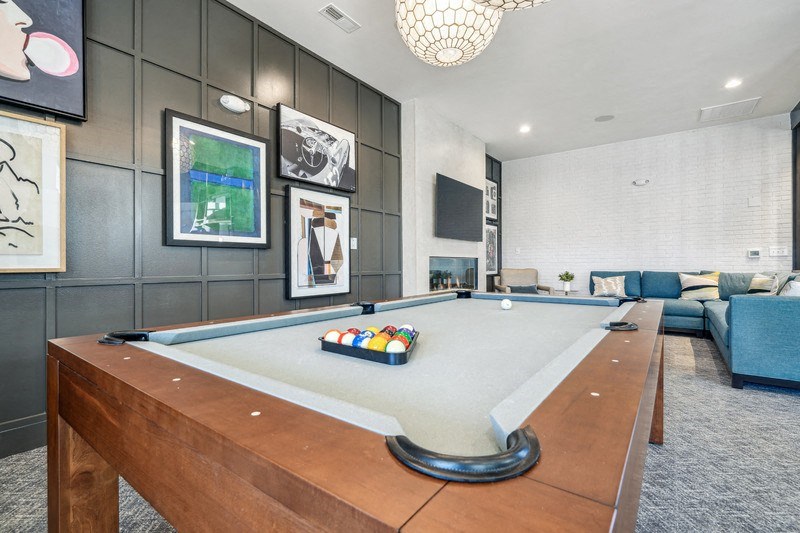 A pool table with a green felt cloth and a white cue ball is in the foreground of a room with a grey carpet.