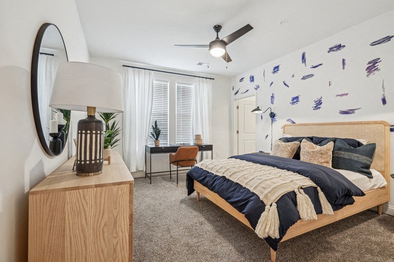 A bedroom with a bed, a chair, a table, a lamp, and a ceiling fan.