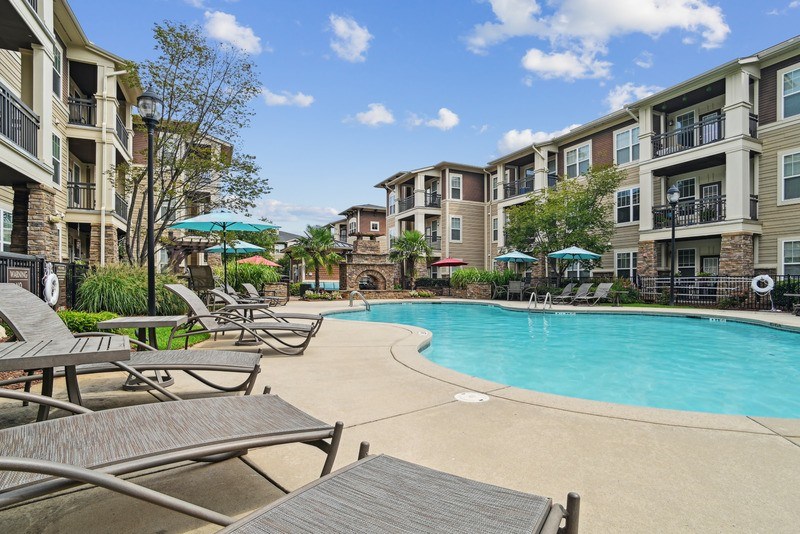 A pool surrounded by lounge chairs and umbrellas in front of apartment buildings.