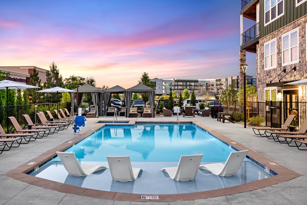 A pool with lounge chairs and umbrellas is surrounded by buildings.