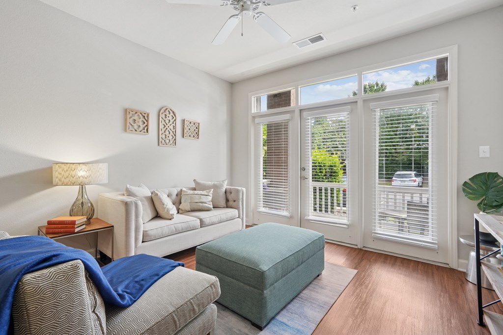 A living room with a grey couch, a green ottoman, and a white table with a lamp on it.