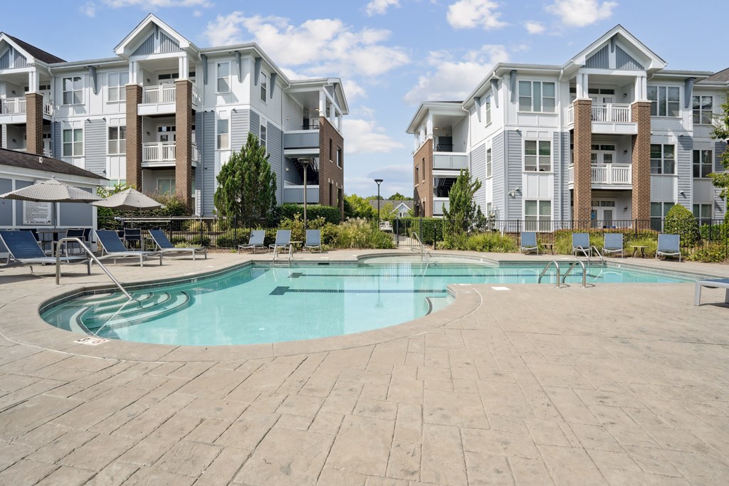 A large swimming pool surrounded by a brick patio and lounge chairs in front of apartment buildings.
