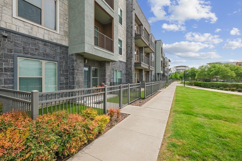 A long concrete walkway leads to a building with a metal fence and bushes on the side.