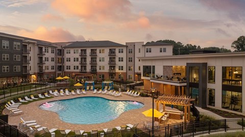 A large swimming pool surrounded by lounge chairs and umbrellas in front of apartment buildings.