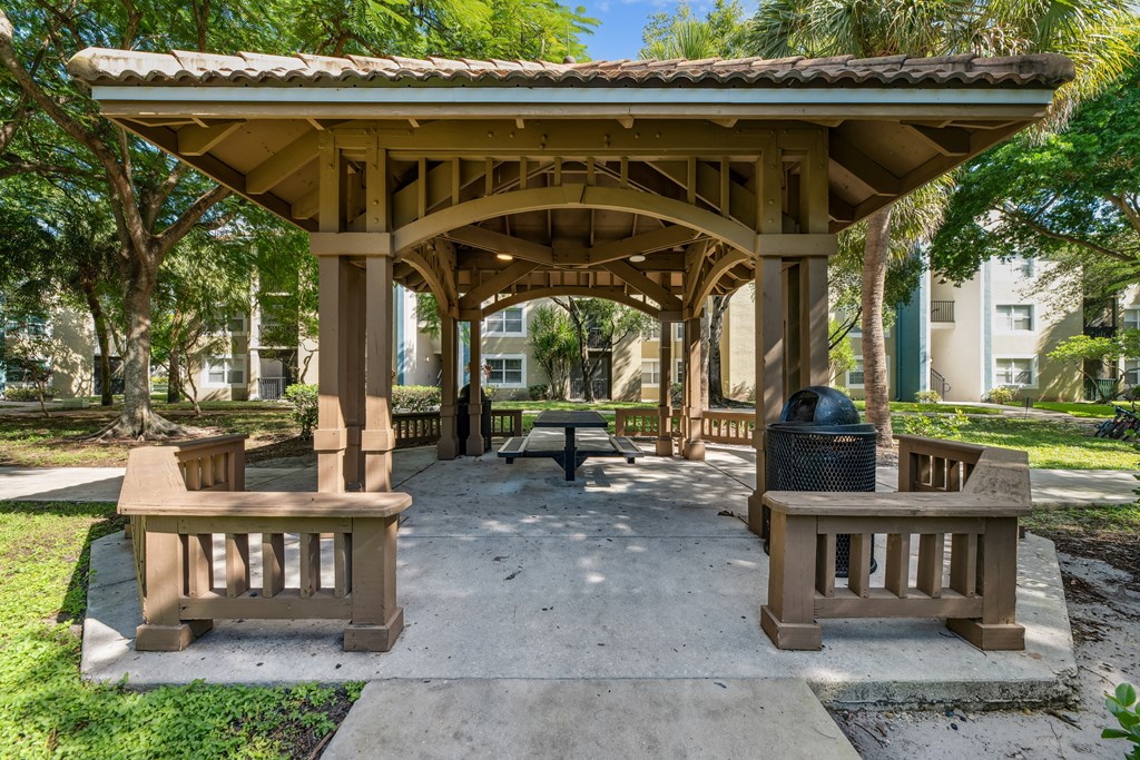 A gazebo with a tiled roof is surrounded by trees and has two benches in front of it.