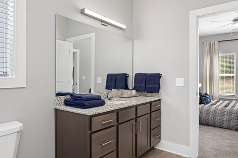 A bathroom with a white toilet, a sink with a mirror, and a bedroom with a bed and a window.
