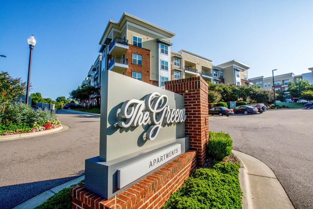 The sign for The Green apartments is in front of a street.