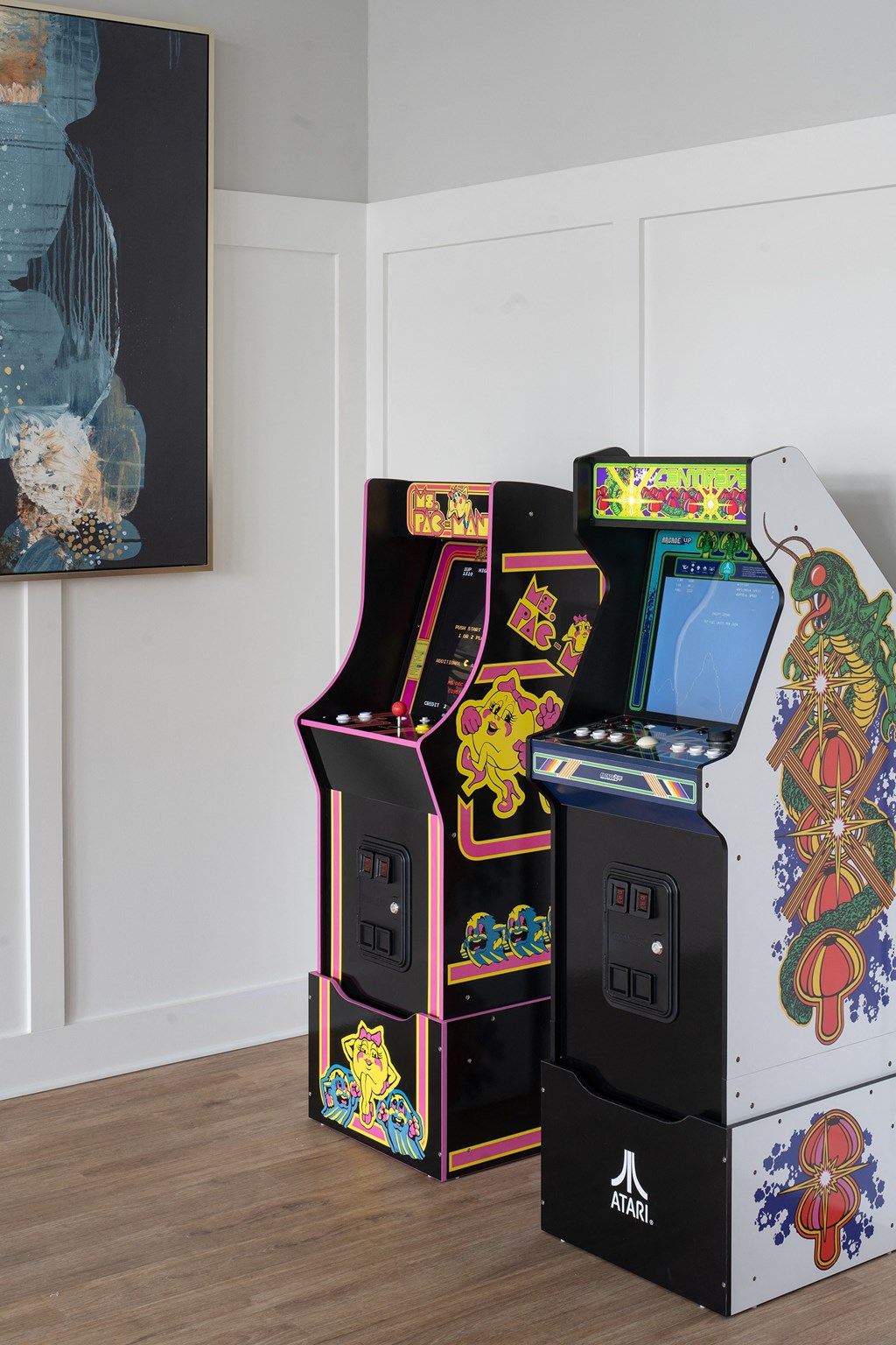 two arcade games in a room with a painting on the wall