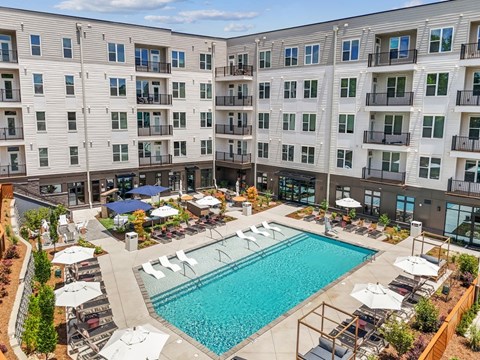 A large swimming pool surrounded by lounge chairs and umbrellas in front of a multi-story apartment building.
