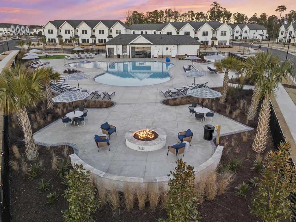 A fire pit is surrounded by chairs and tables in a courtyard with a pool behind it.