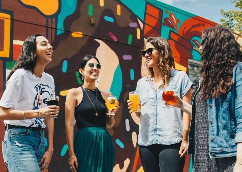 a group of women standing in front of a mural holding drinks