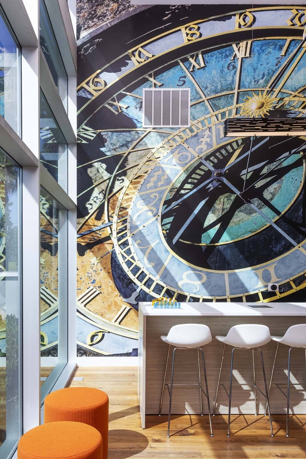 A large mural of a clock is on the wall of a room with a bar and stools.