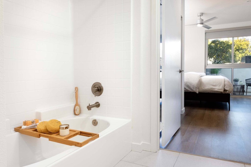 A white bathroom with a tub and a bedroom in the background.
