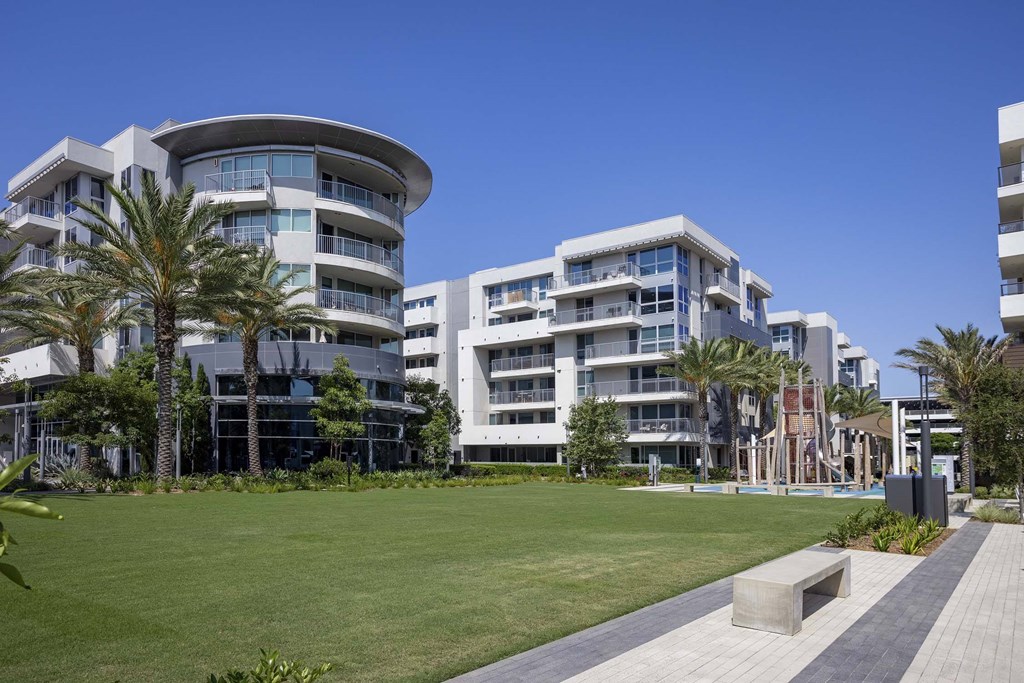 A modern building with a curved design and balconies is surrounded by a grassy area and palm trees.