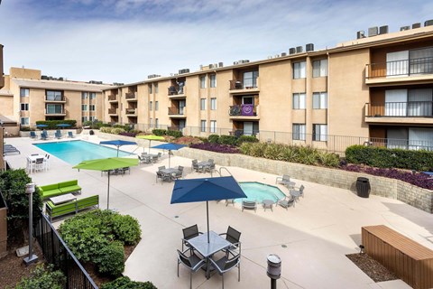 A large outdoor pool area with lounge chairs and umbrellas in front of apartment buildings.
