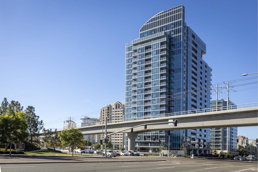 A modern glass building with a curved top is situated in a parking lot with a bridge overhead. at LUX by Garden*, San Diego