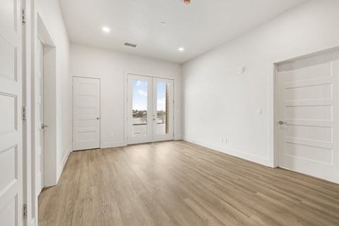 A room with white walls and wooden flooring, with a view of a building outside the window.