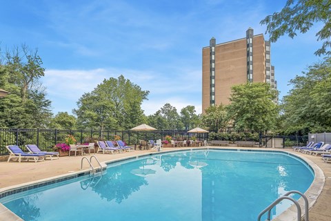 A large swimming pool surrounded by trees and chairs with a tall building in the background.