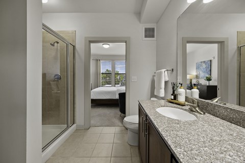 A bathroom with a sink, mirror, toilet, and a bedroom with a bed and window.