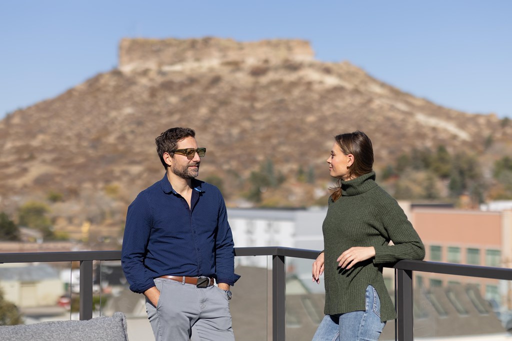 A man and a woman are standing on a balcony with a large hill in the background.