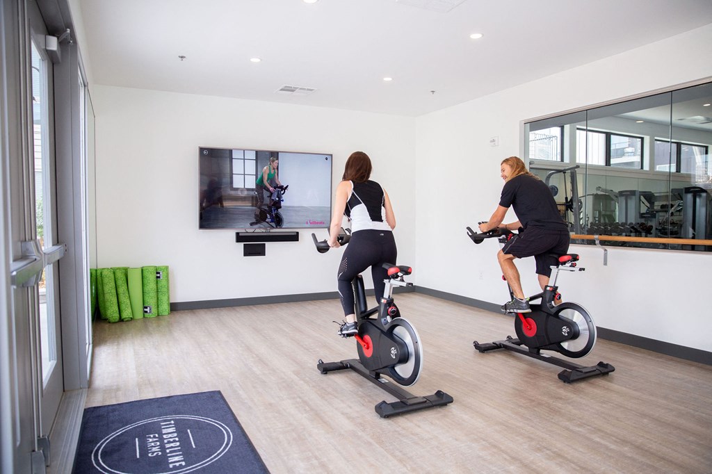 Two people are exercising on stationary bikes in a well-lit room.