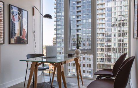 A modern dining room with a glass table and chairs, a laptop, a plant, and a framed picture on the wall.