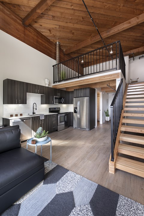 A black leather couch sits in a living room with a wooden floor and a staircase.