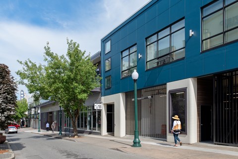 A woman in a straw hat walks down a sidewalk past a blue building at B Flats Apartments, Bremerton, 98337
