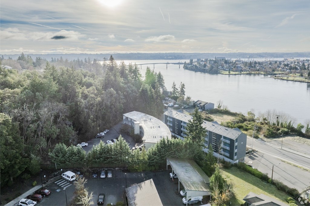 A sunny day at a parking lot with a large building and a body of water in the background.