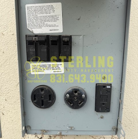 A grey electrical box with a warning label and a yellow label with the text "STERLING" and a phone number.
