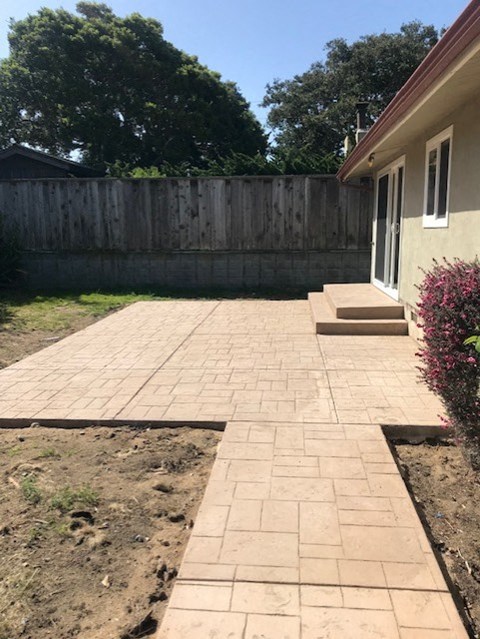 A patio with a brick pattern is in front of a house and a wooden fence.