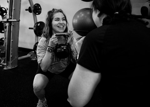 A woman is squatting in a gym with a personal trainer standing next to her.