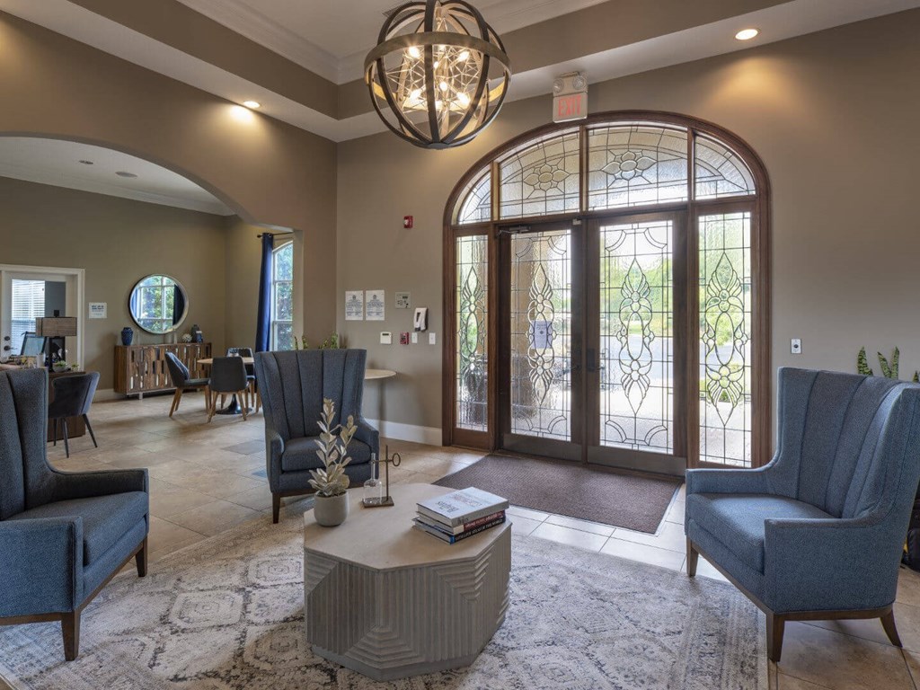 A living room with a grey sofa, a grey armchair, a grey coffee table, a chandelier, and a large window with a view of trees.
