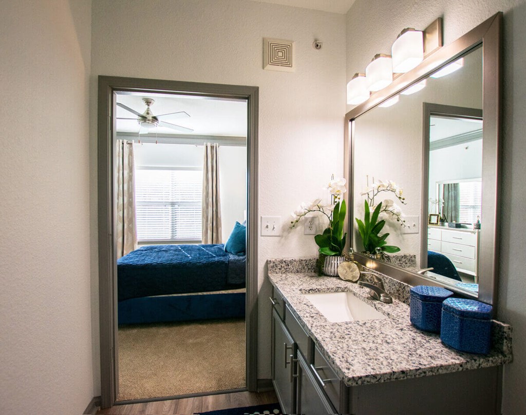 A bathroom with a mirror, sink, and a bedroom with a bed and a window.