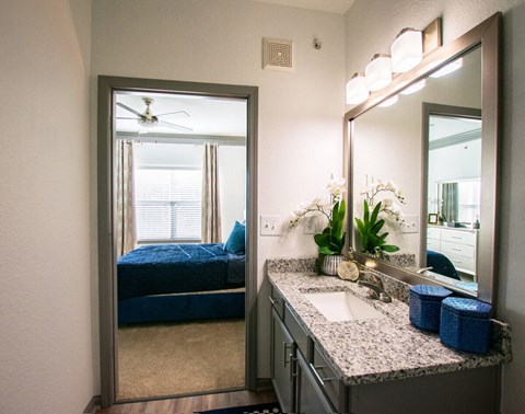 A bathroom with a mirror, sink, and a bedroom with a bed and a window.