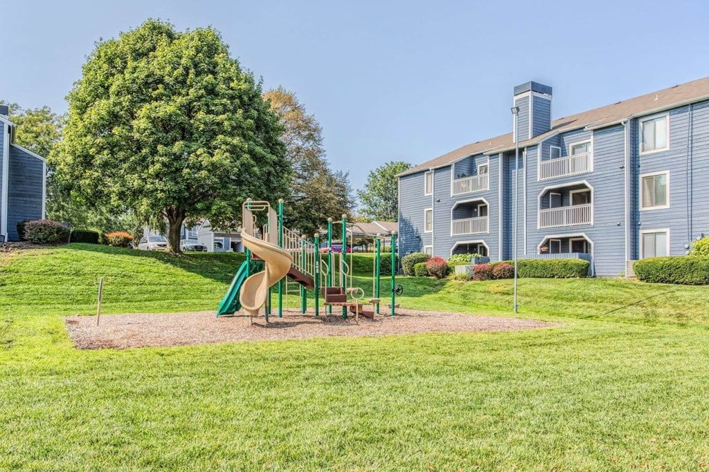 A playground with a slide and a green tree in front of a building.