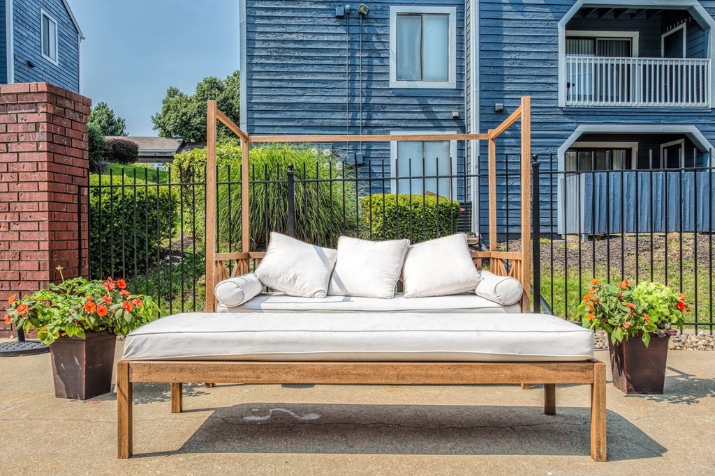 A wooden daybed with a white cushion and two pillows is placed in front of a blue building with a black fence.