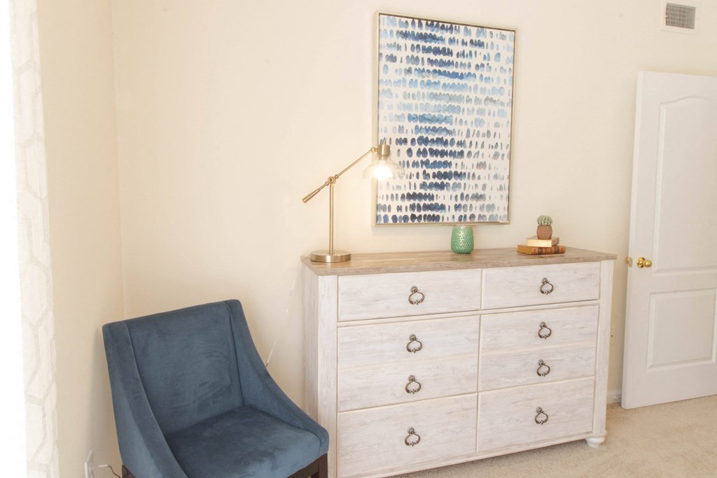 A blue chair sits in front of a white dresser with a framed artwork on the wall. at The Crest at Sugarloaf, Lawrenceville, Georgia