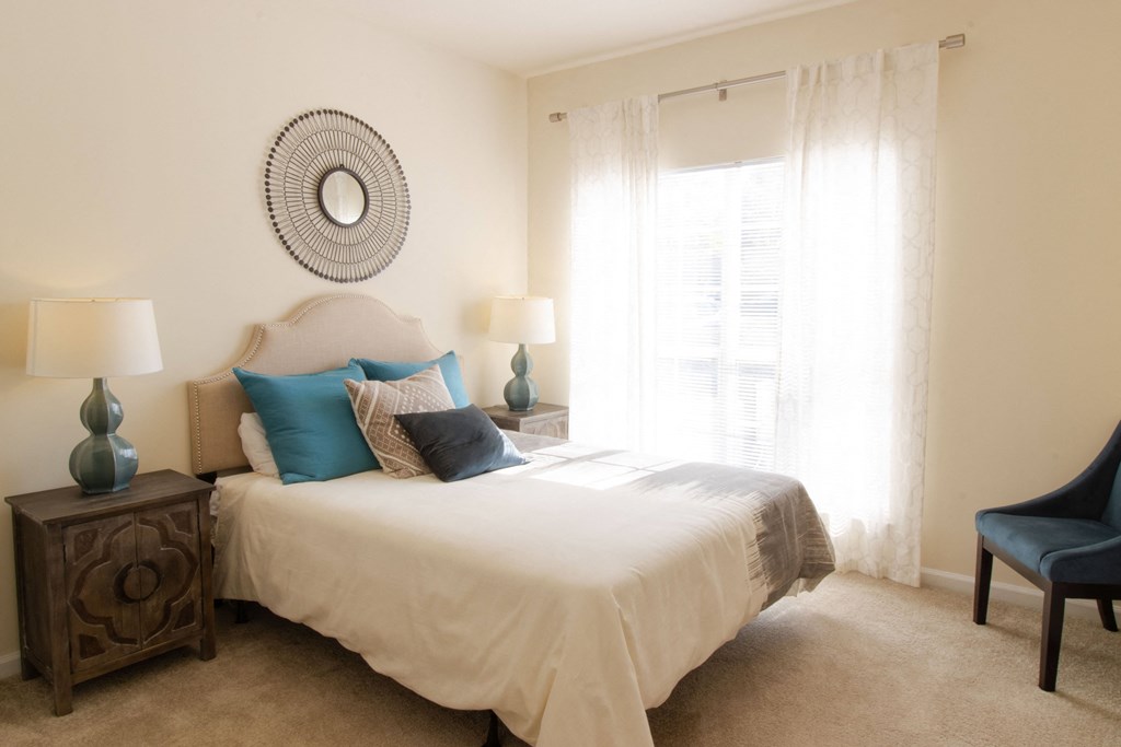 A bedroom with a bed, two lamps, a chair, and a mirror on the wall. at The Crest at Sugarloaf, Lawrenceville