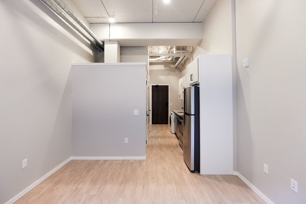 an empty room with white walls and a wood floor and a kitchen