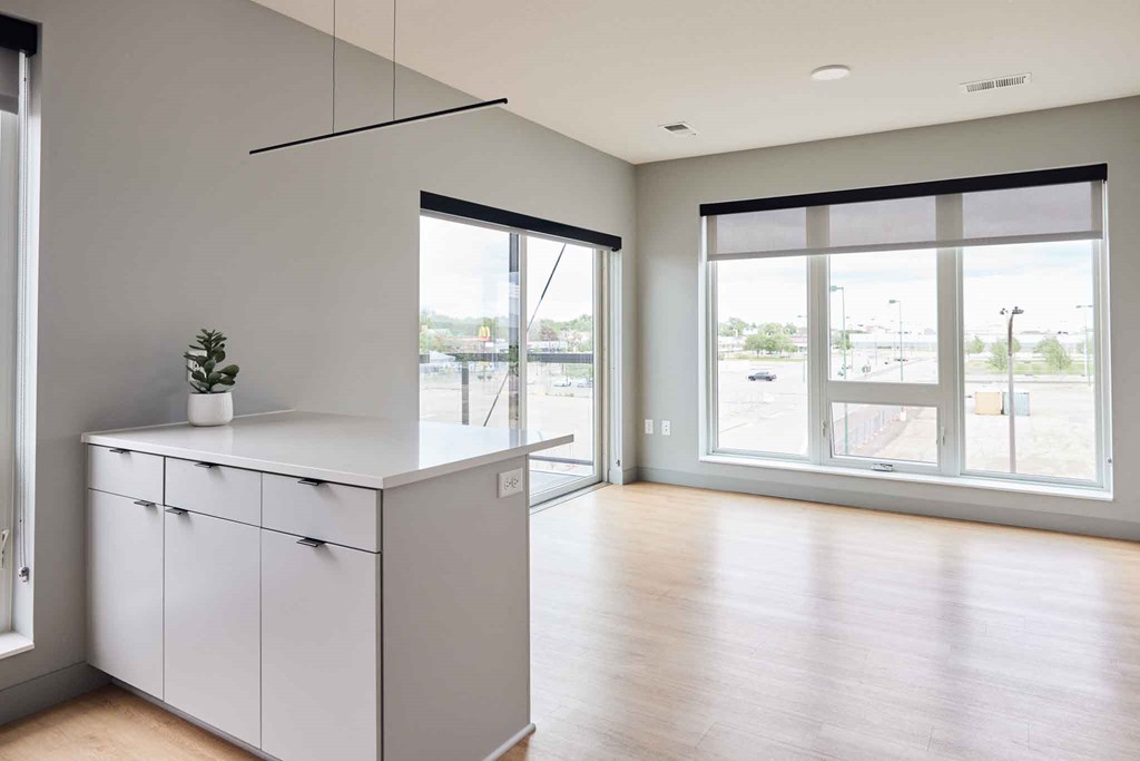 an empty living room with a large window and a kitchen with white cabinets