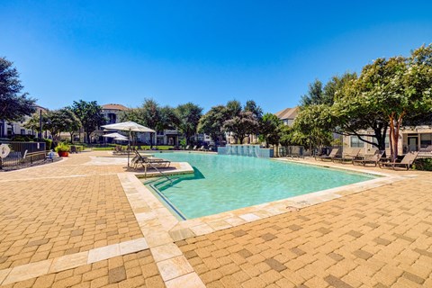 A large outdoor swimming pool surrounded by a brick patio and lounge chairs.