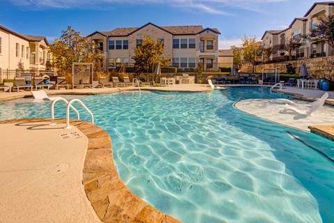A large swimming pool with a sandy edge and a residential building in the background.