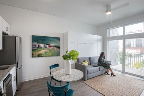 A woman is sitting on a grey couch in a living room with a painting on the wall.