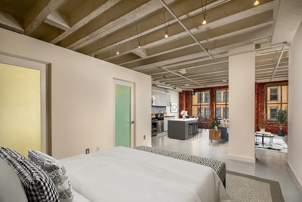 A bedroom with a bed and a kitchen area in the background.