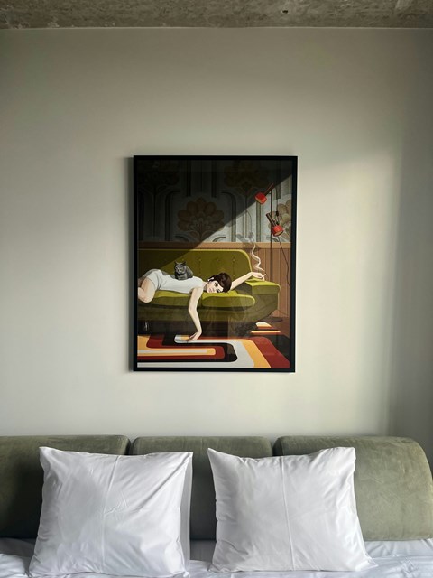 A painting of a cat on a couch is hanging on the wall above a bed with two pillows.