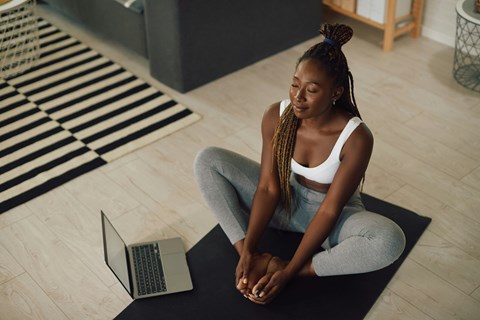 A woman is sitting cross-legged on a mat with a laptop on her lap.