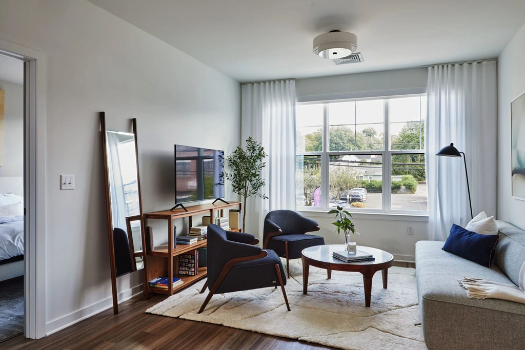 A living room with a grey couch, a wooden coffee table, a chair, a desk with a computer, a potted plant, and a large window with white curtains.