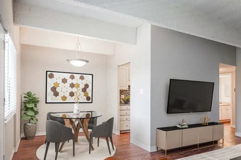 A modern dining room with a glass table and chairs, a framed artwork on the wall, and a television on a cabinet.